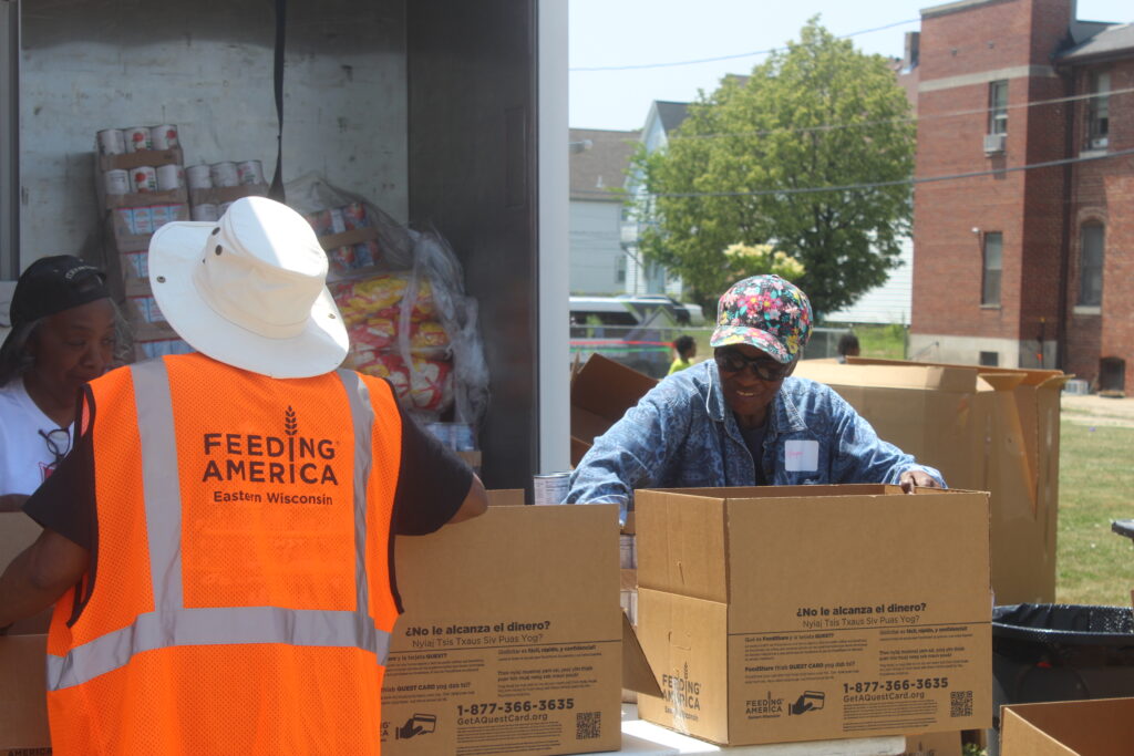 Volunteers in orange vests pack food items into boxes, with a truck filled with bags of food behind them.