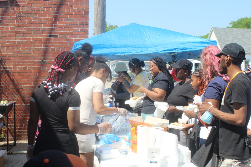 Community members enjoying food under a blue canopy at an outdoor event.